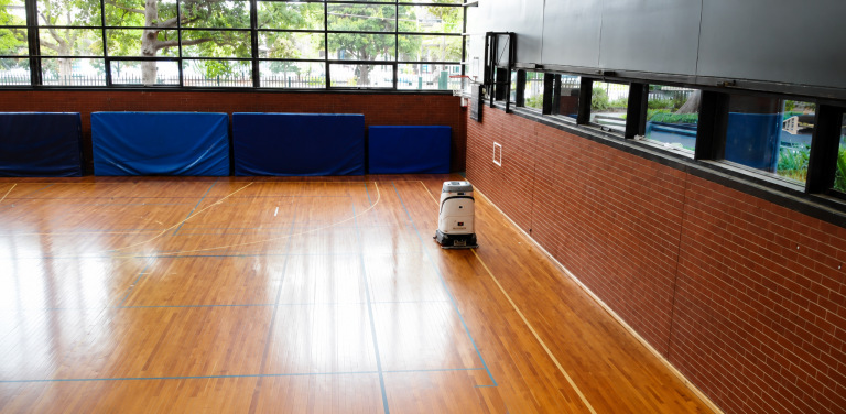 Quad 800 autonomous floor cleaning robot cleaning a polished indoor basketball court in a sports facility with timber flooring and court line markings.